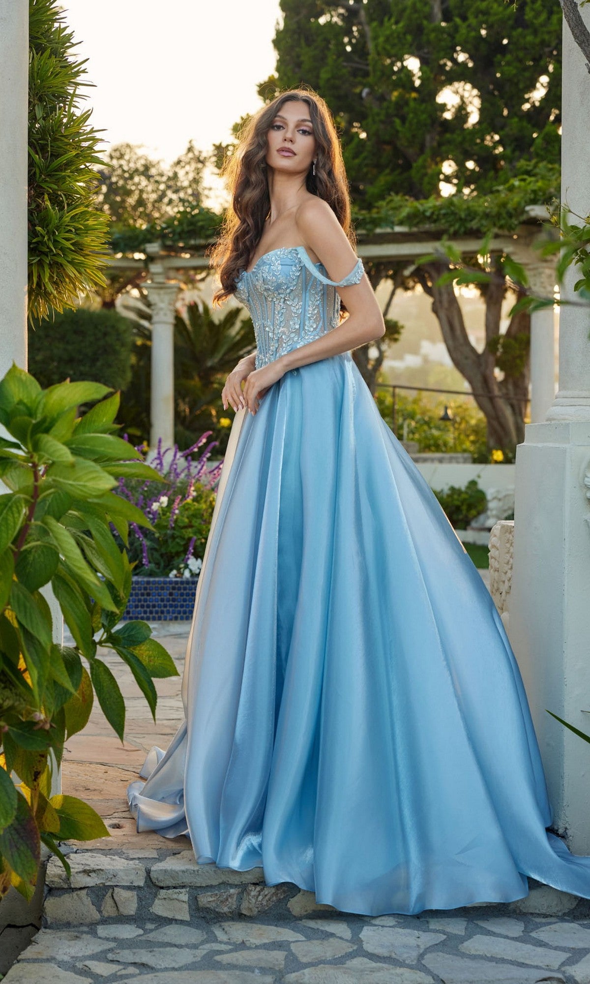 A woman stands outdoors in the Juliet JT2639G, a light blue off-the-shoulder A-line satin prom dress with a fitted corset bodice and full skirt, surrounded by greenery and stone structures.